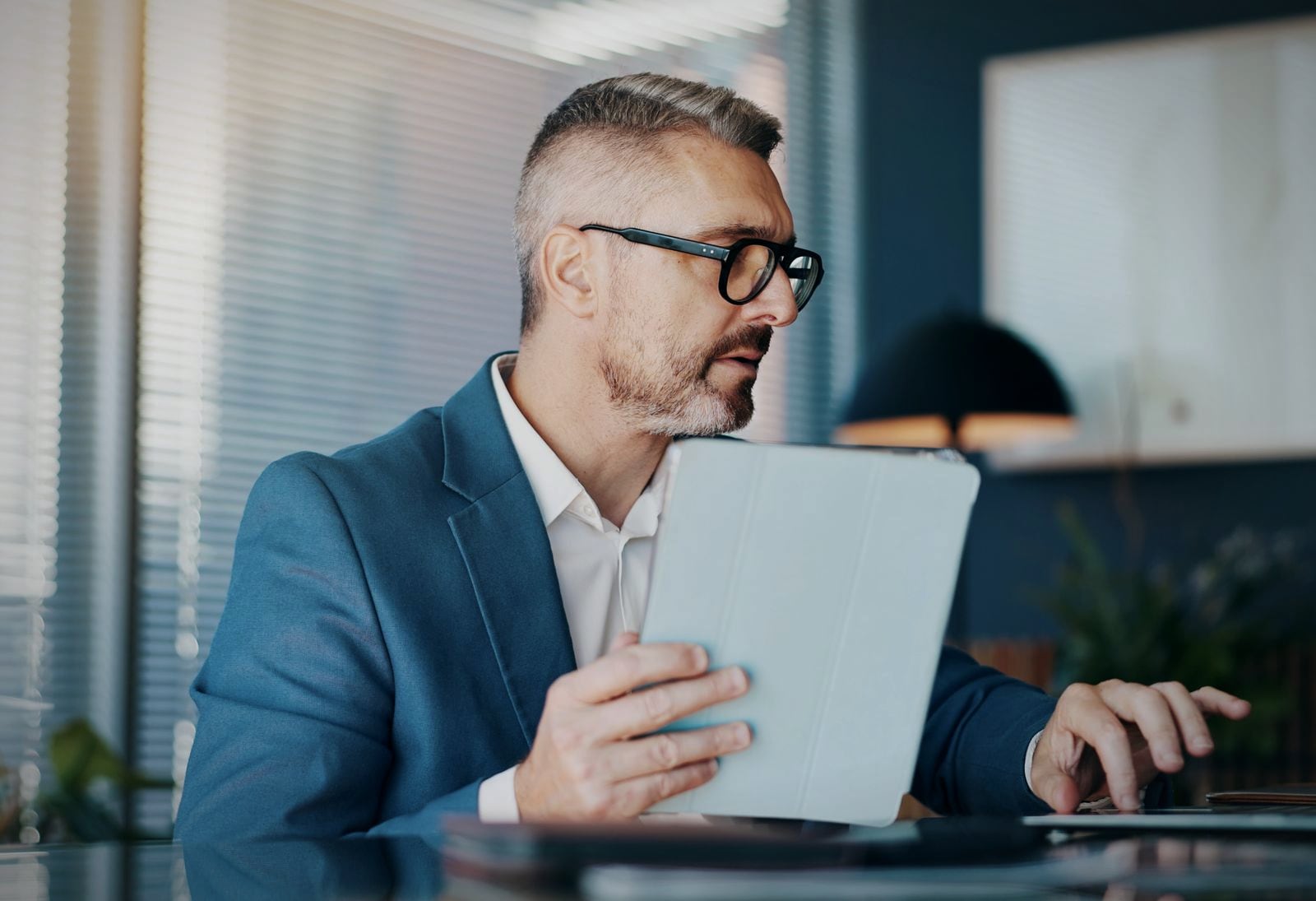 A manager at an office desk with a tablet and laptop, looking up a figure in the company's own data